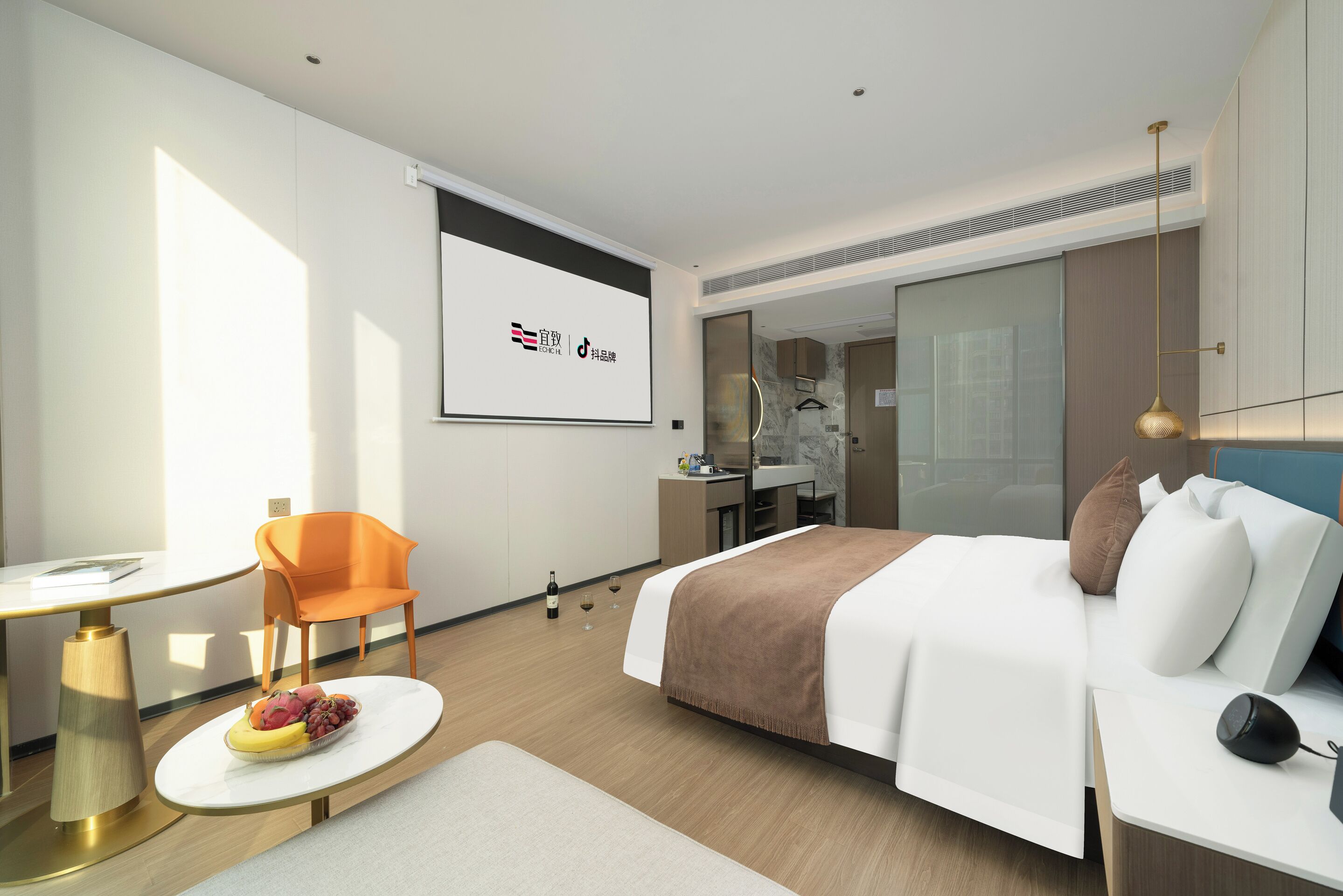 A modern hotel room with a bed, a desk, a chair, and a small table with fruit.
