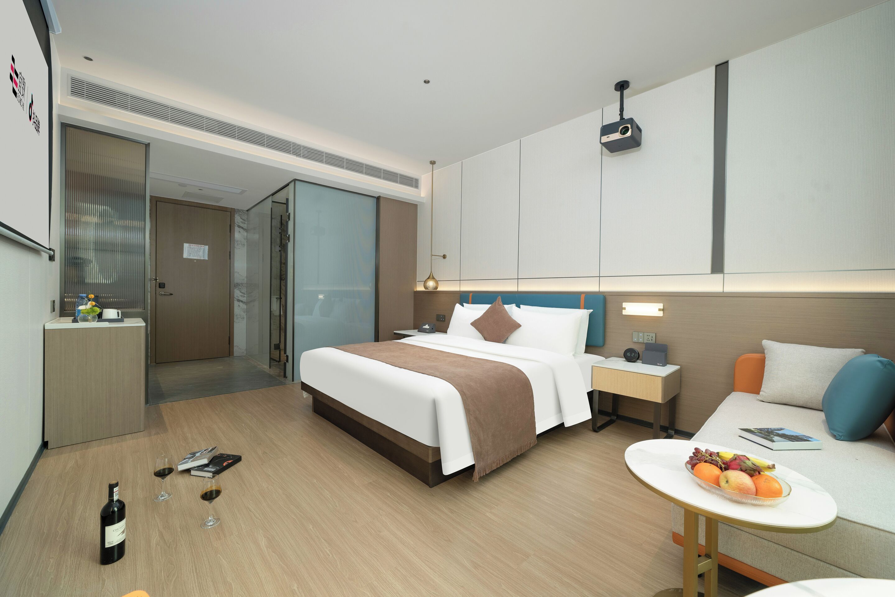 Ein modernes Hotelzimmer mit einem großen Bett, Nachttischen, einem kleinen Schreibtisch, einer Sitzecke mit einer Couch und einem Esstisch mit Obst.