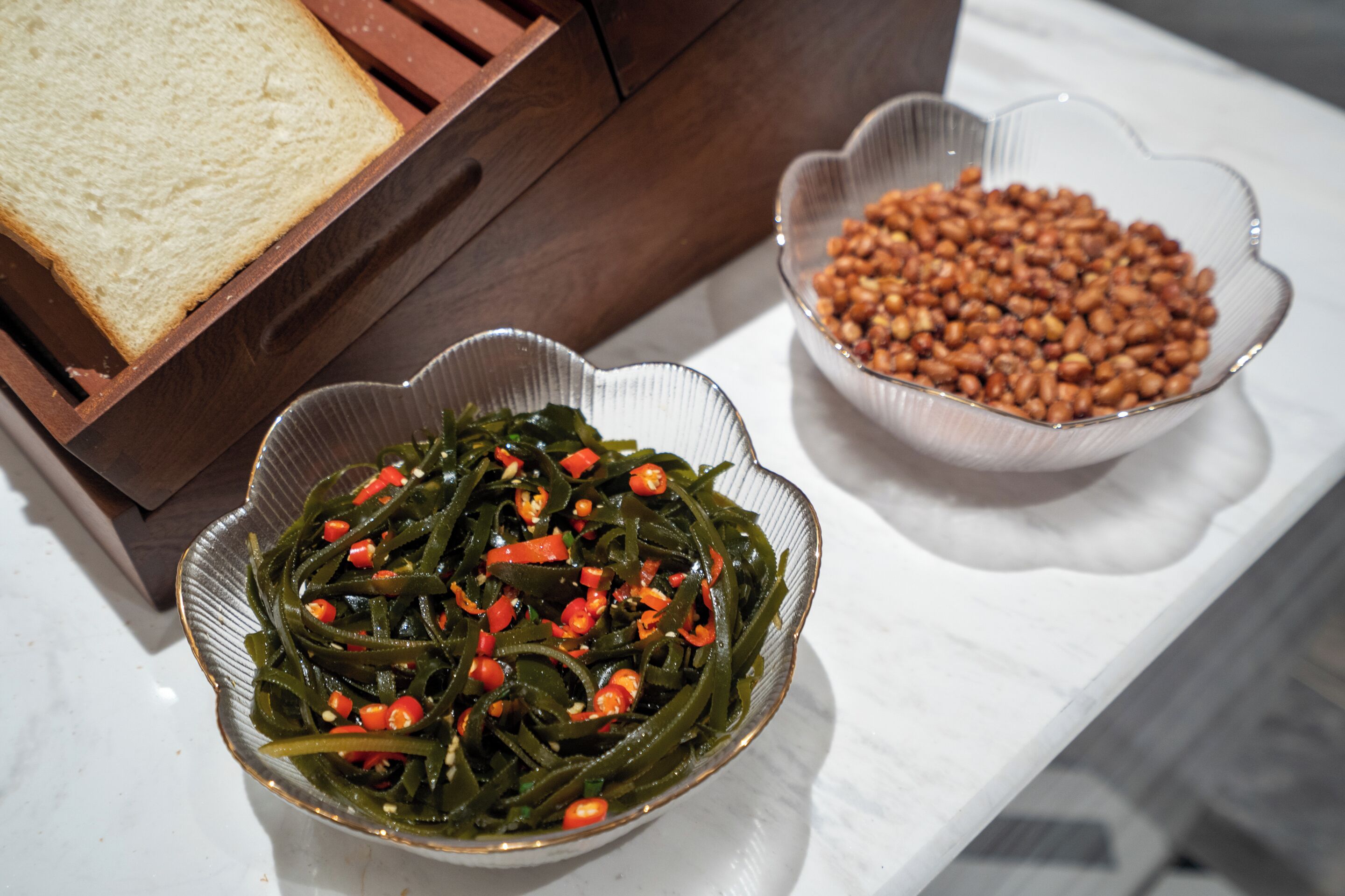 A table with a bowl of green vegetables, a bowl of peanuts, and a slice of bread.