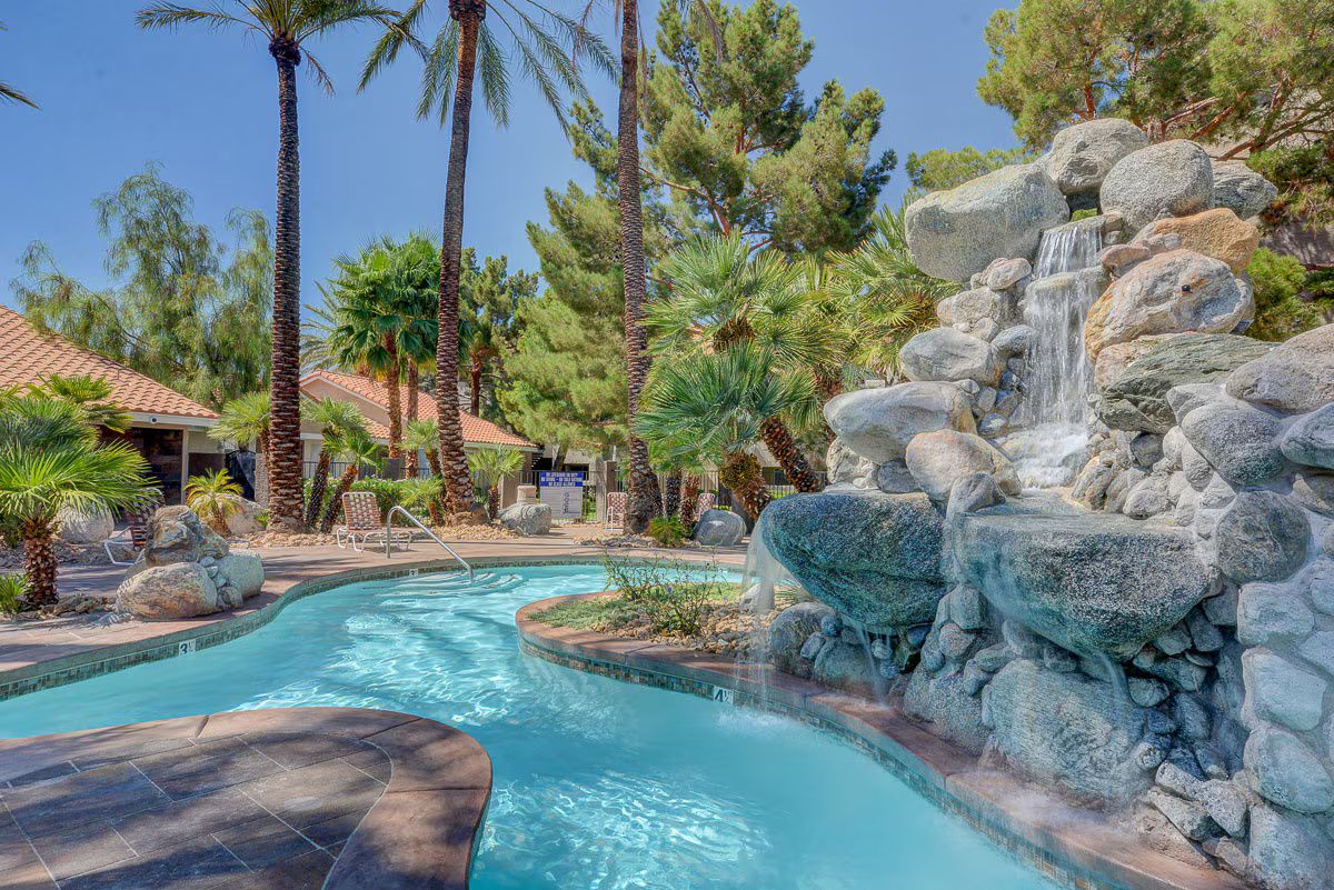 Zona de piscina amb cascada, rodejada de palmeres i un edifici amb teulada enrajolada.