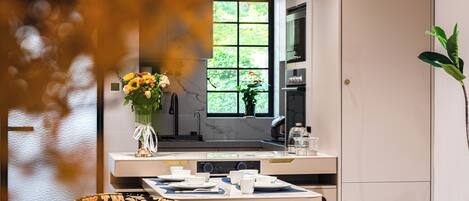 A modern kitchen with a dining area, a window with a view of greenery, and a vase with flowers on the counter.