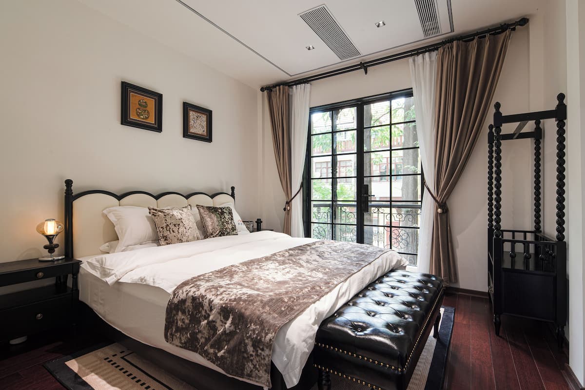 A neatly arranged bedroom with a large bed, a bedside table with a lamp, a window with curtains, and a black wardrobe.