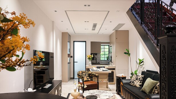 Sala de estar moderna com sofá, mesa de centro e televisão. Escada e cozinha ao fundo.