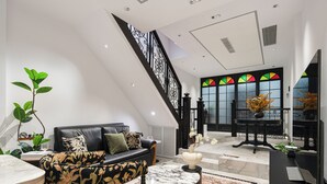 Uma sala de estar moderna com um sofá de couro preto, uma poltrona estampada, uma mesa de centro de mármore e uma escada com corrimãos pretos.