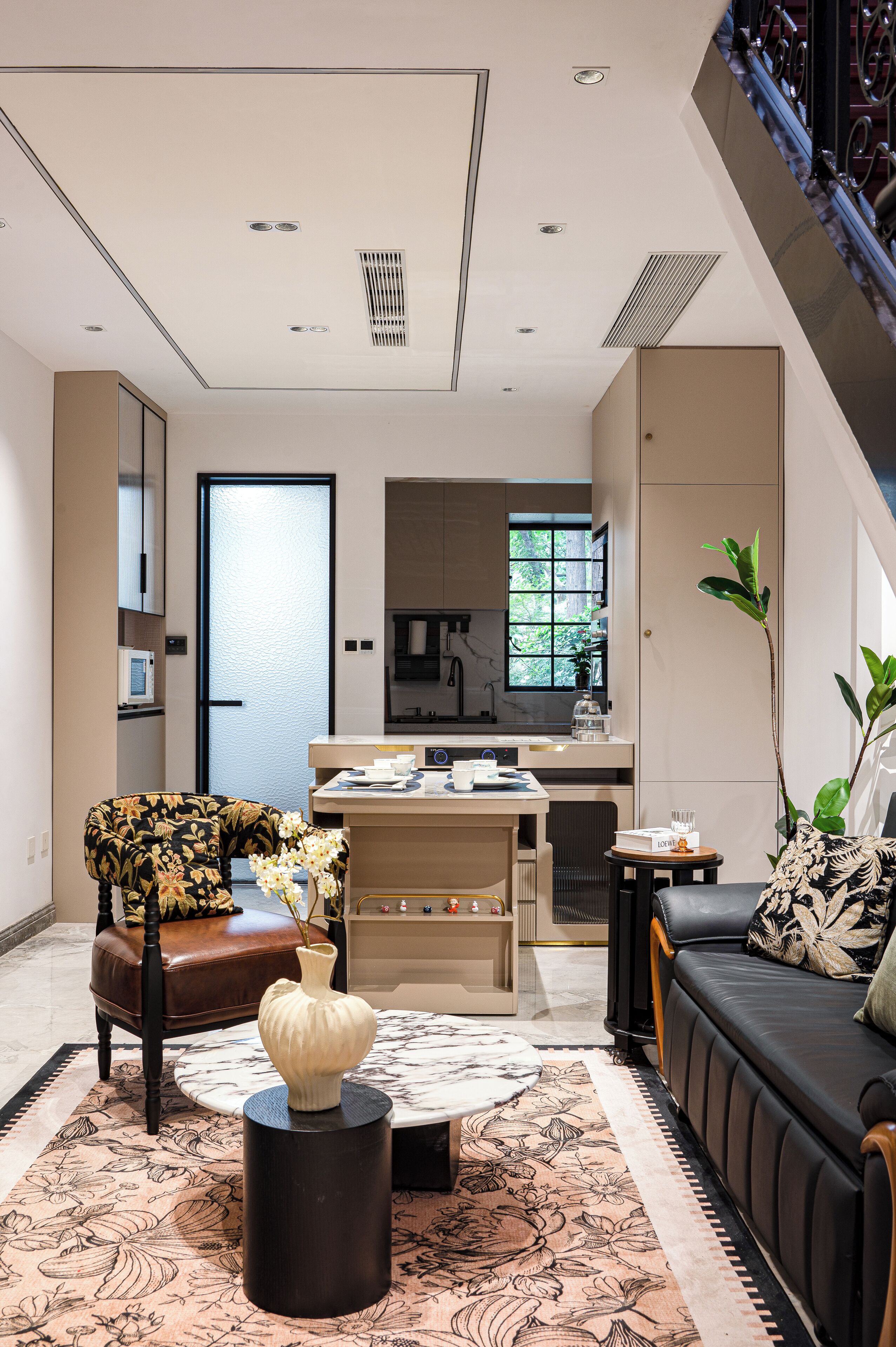 Una sala moderna con un sofá, una mesa de centro y una silla. Se observa una cocina al fondo, con una ventana y un microondas.