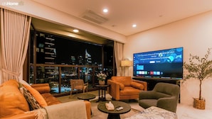 A modern living room with a large TV, comfortable seating, a view of the city at night, and a potted plant.