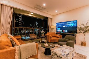 A modern living room with a large TV, comfortable seating, a view of the city at night, and a potted plant. - Gallery Residence - Metropole Thu Thiem (Ho Chi Minh)