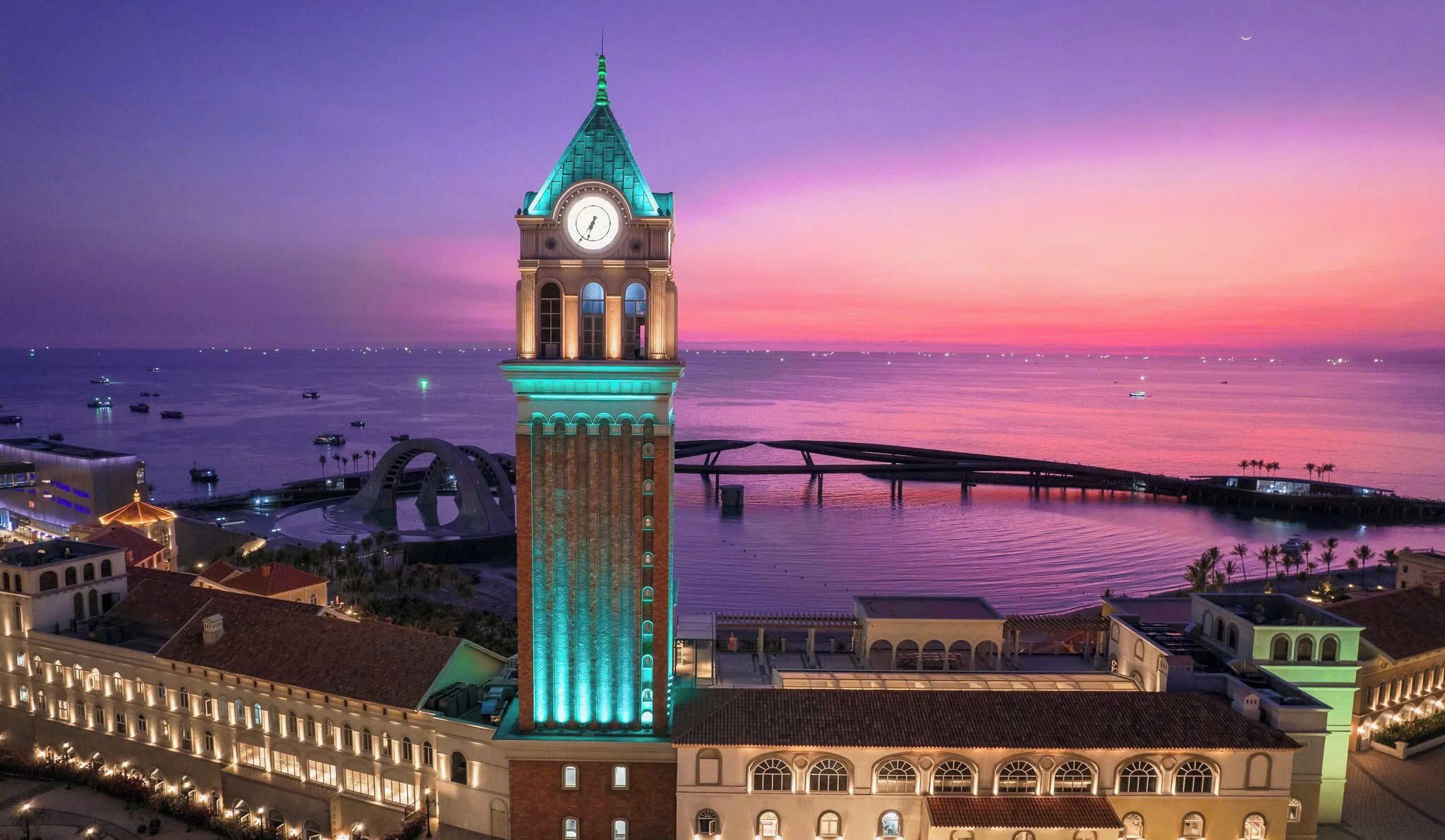 Uma torre de relógio histórica com mostrador, rodeada por edifícios e uma orla marítima.