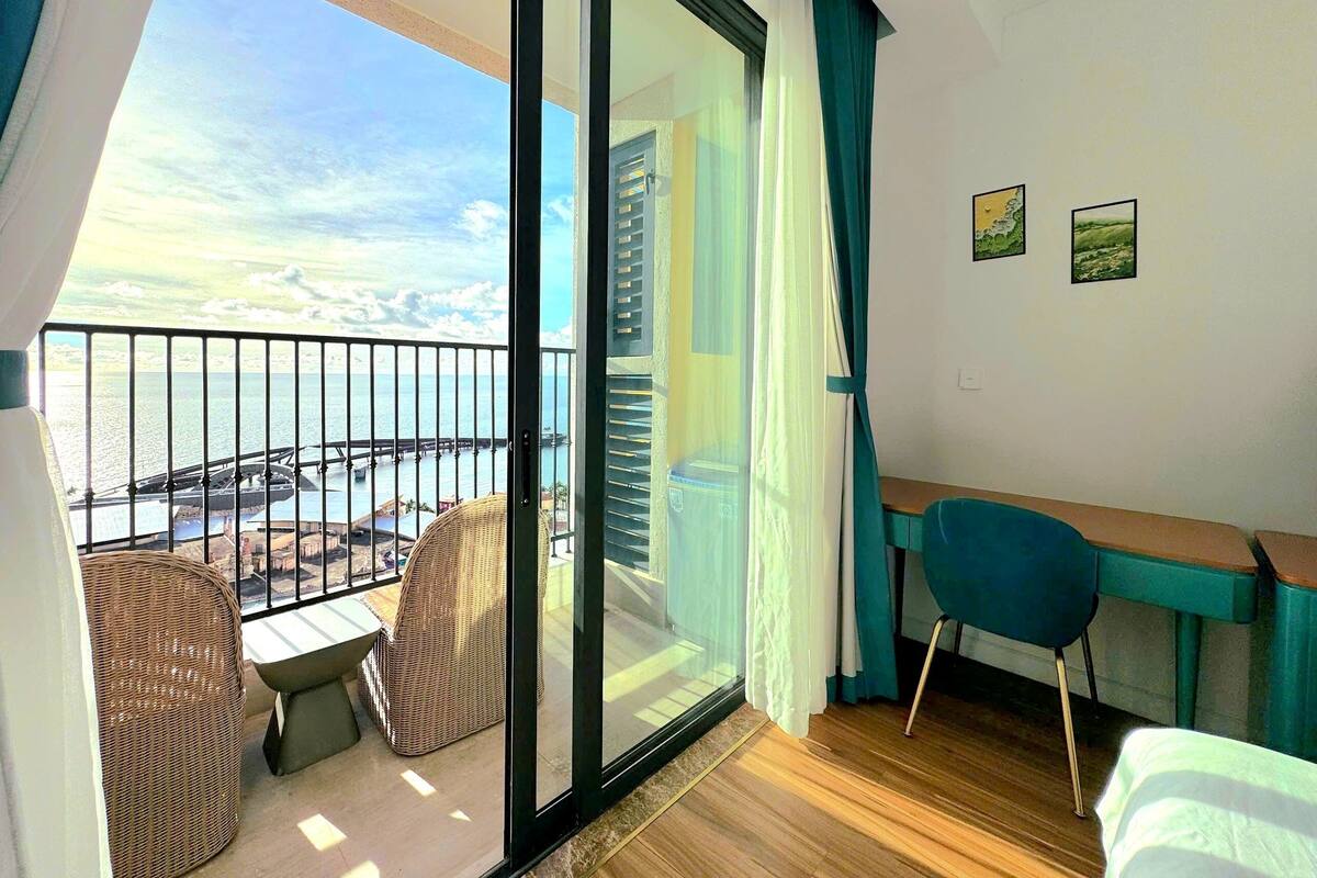 A balcony with a view of a bridge and water, a wicker chair, a desk with a chair, and a bed.