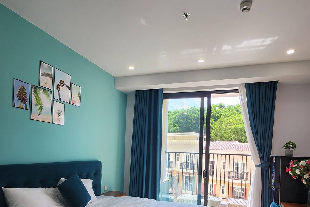 A bedroom with a bed, a view to the outside, a ceiling light, and a wall with pictures.