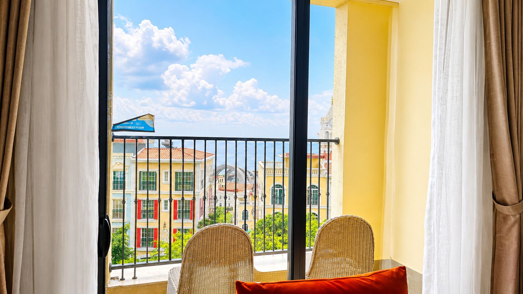 A balcony with wicker chairs and a red cushion, overlooking a building with red shutters and a blue awning.