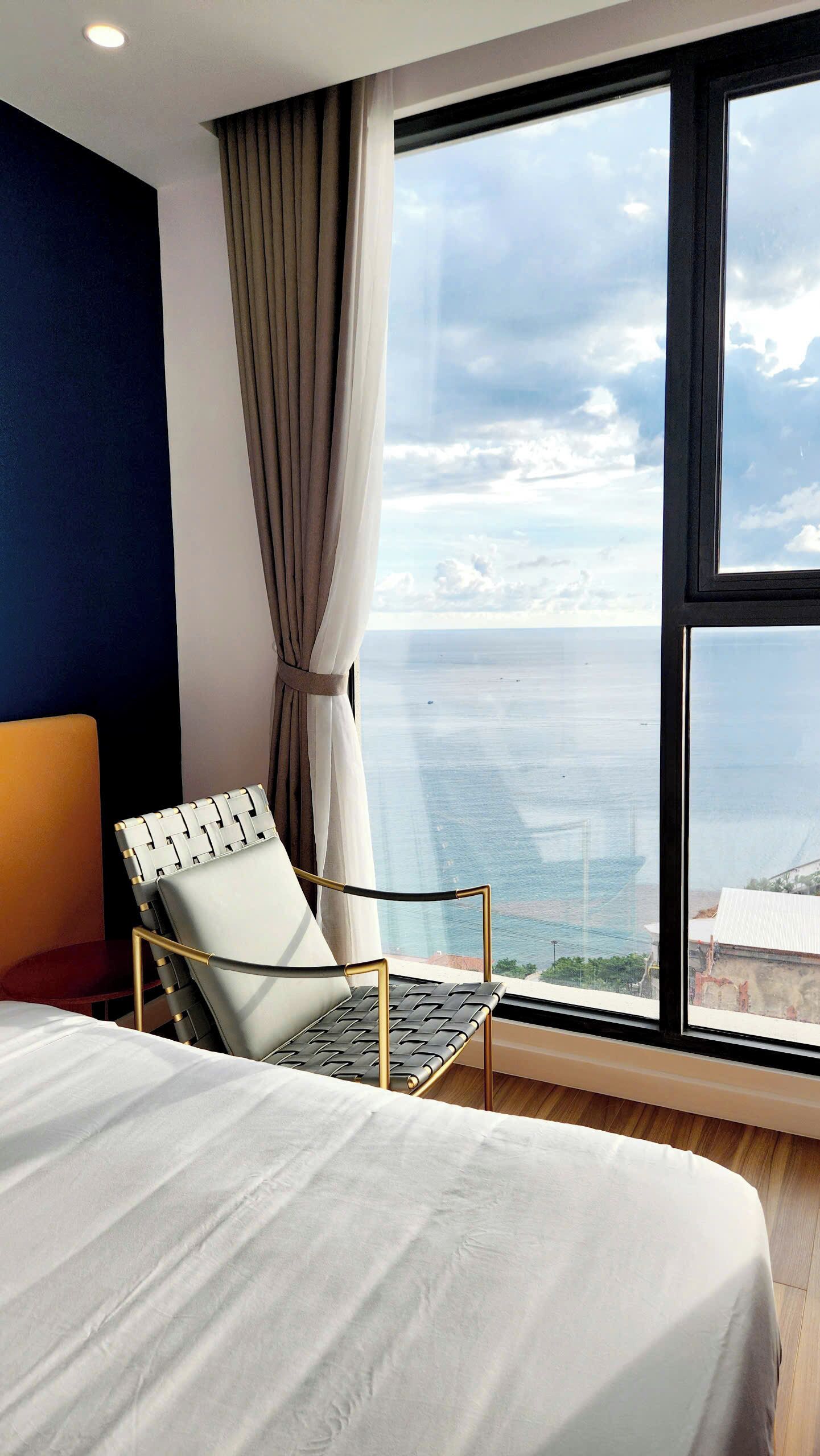 Una moderna camera d'hotel con una grande finestra vista mare, un letto con lenzuola bianche, una sedia nera e dorata e una vista sull'oceano.