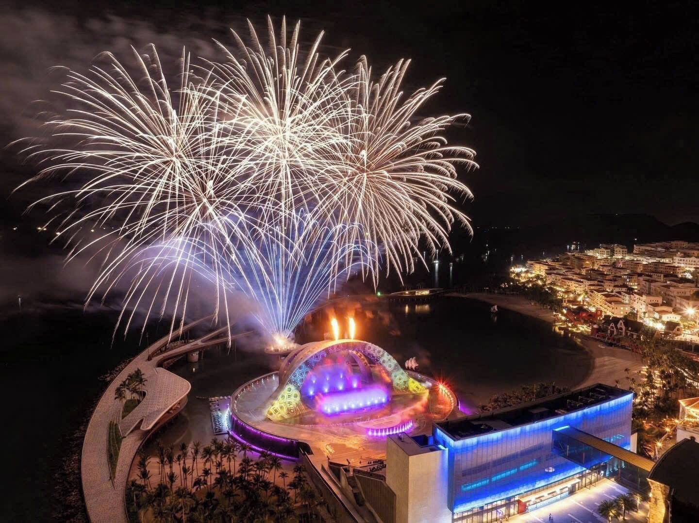 Vista noturna de uma cidade costeira com um espetáculo de fogos de artifício, uma grande estrutura em forma de cúpula iluminada e um edifício moderno com iluminação azul.