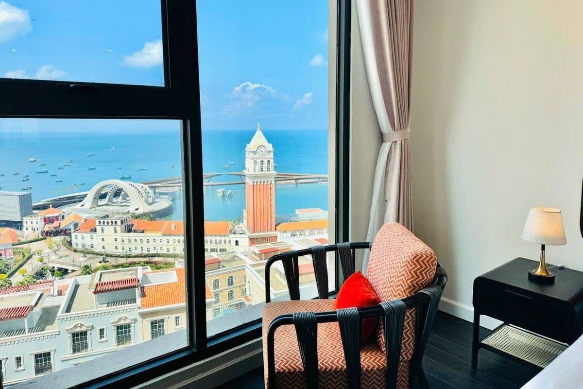 A hotel room with a large window offering a view of a cityscape, including a clock tower and a bridge.