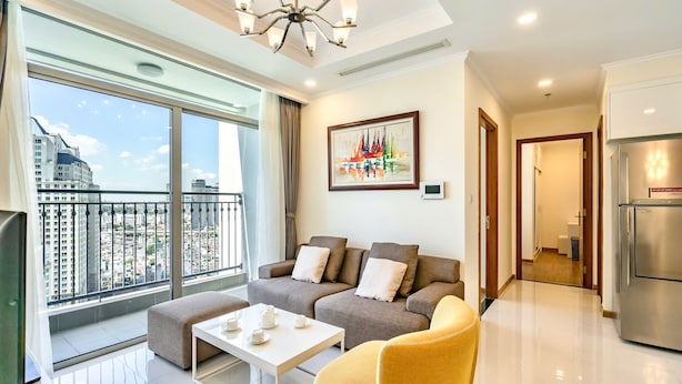 A modern living room with a sofa, ottoman, and coffee table, a large window with a city view, and a painting on the wall.