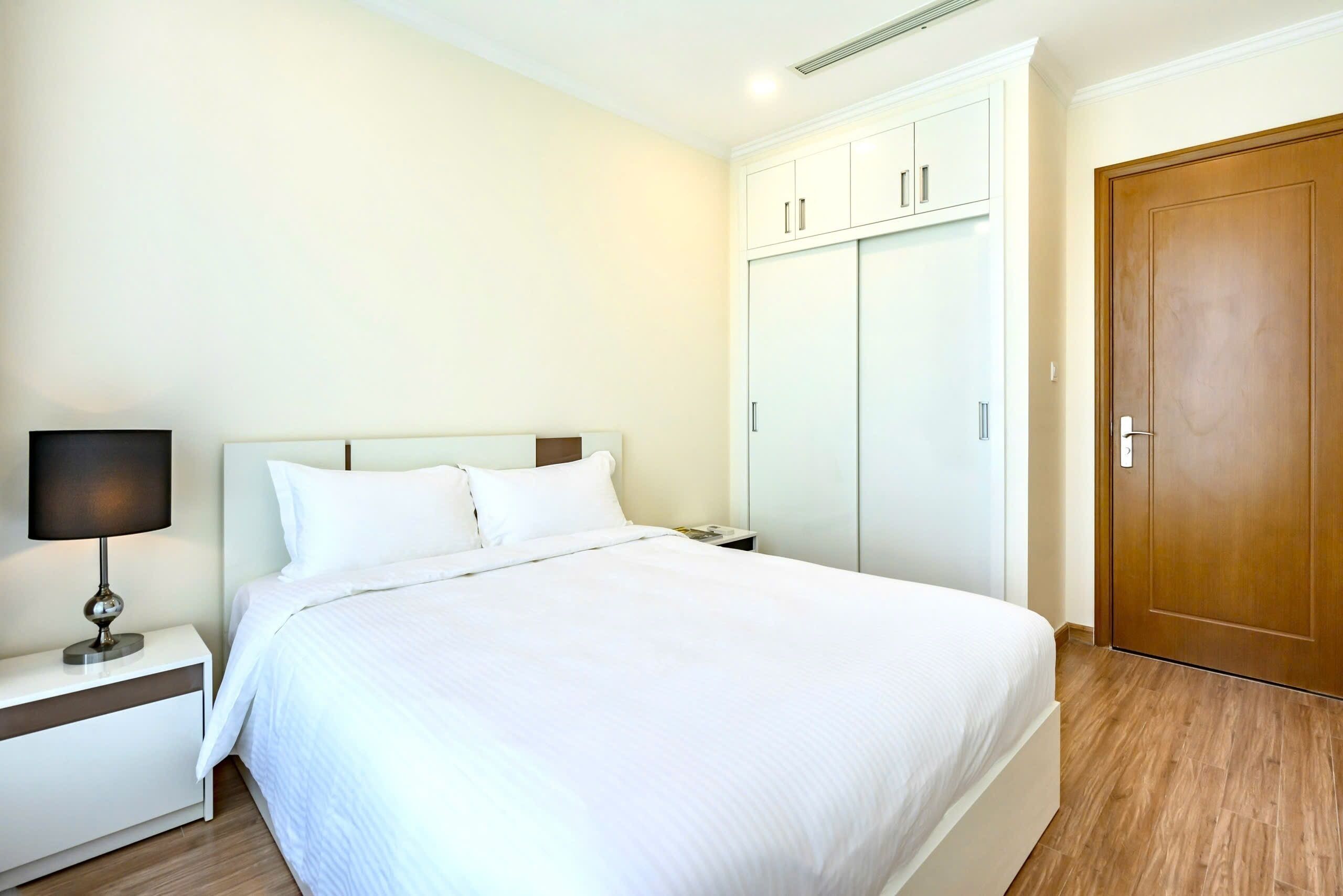 Uma cama bem arrumada com lençóis brancos, uma mesa de cabeceira com abajur, um guarda-roupa com portas deslizantes e uma porta de madeira.
