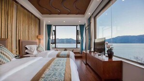 A hotel room with two beds, a TV, and a large window offering a view of a lake and mountains.
