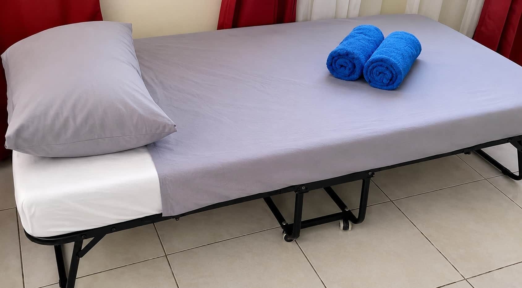 Una cama individual con colchón gris, almohada blanca y dos toallas enrolladas en azul.