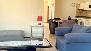 Un salon comprenant un canapé, une table basse ornée d’un chemin de table en dentelle, une lampe rouge posée sur une petite table d’appoint en verre, et un coin repas avec une table et des chaises.
