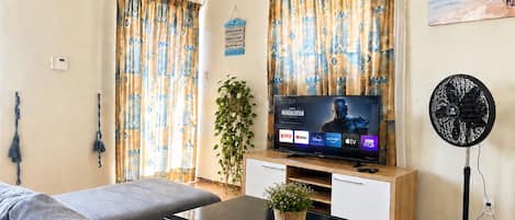 A living room with a flat-screen TV displaying 'Mandalorian', a sofa, a coffee table, a fan, and a plant.