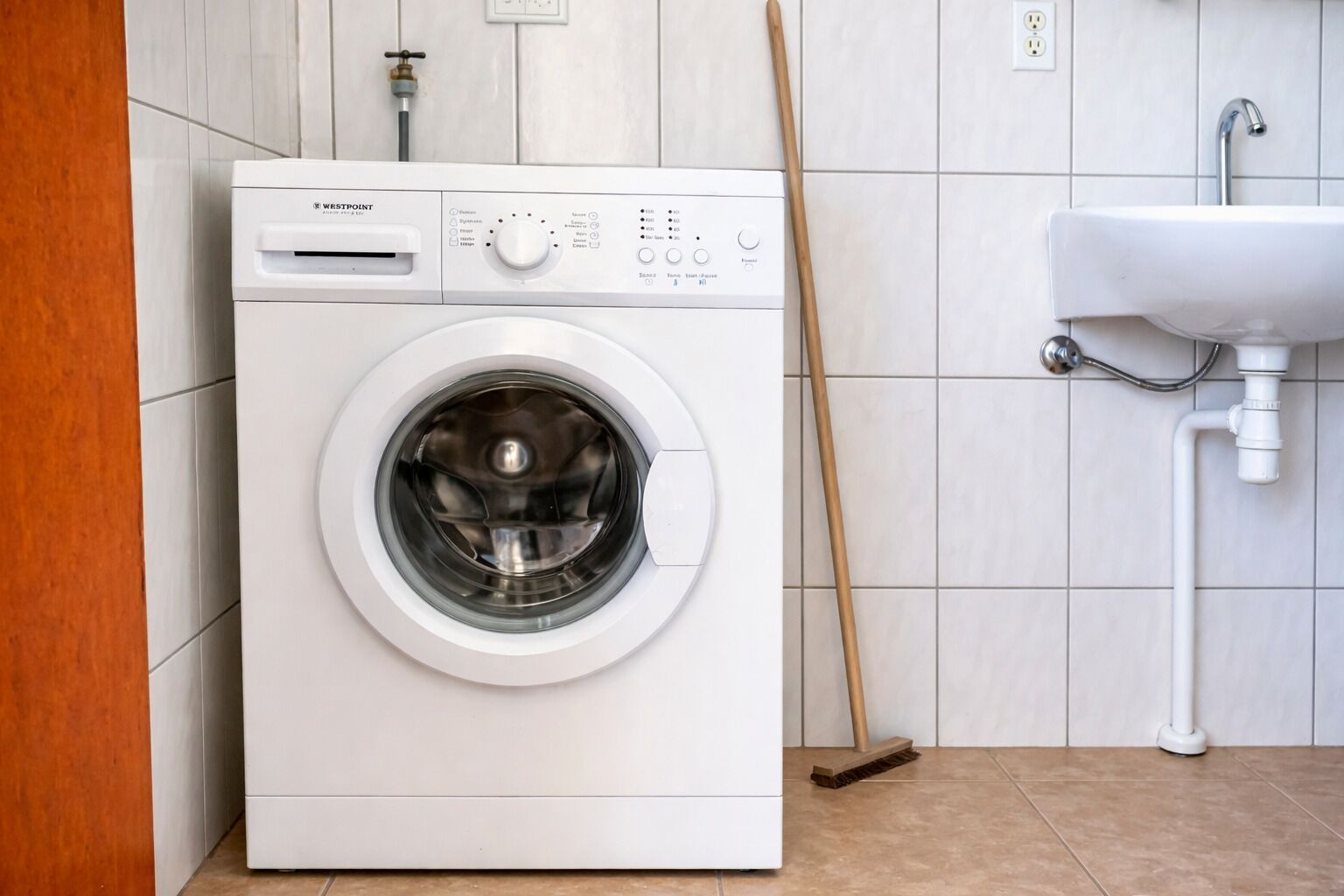 Una m àquina de rentar amb tapa frontal en un bany amb un lavabo i una escombra.