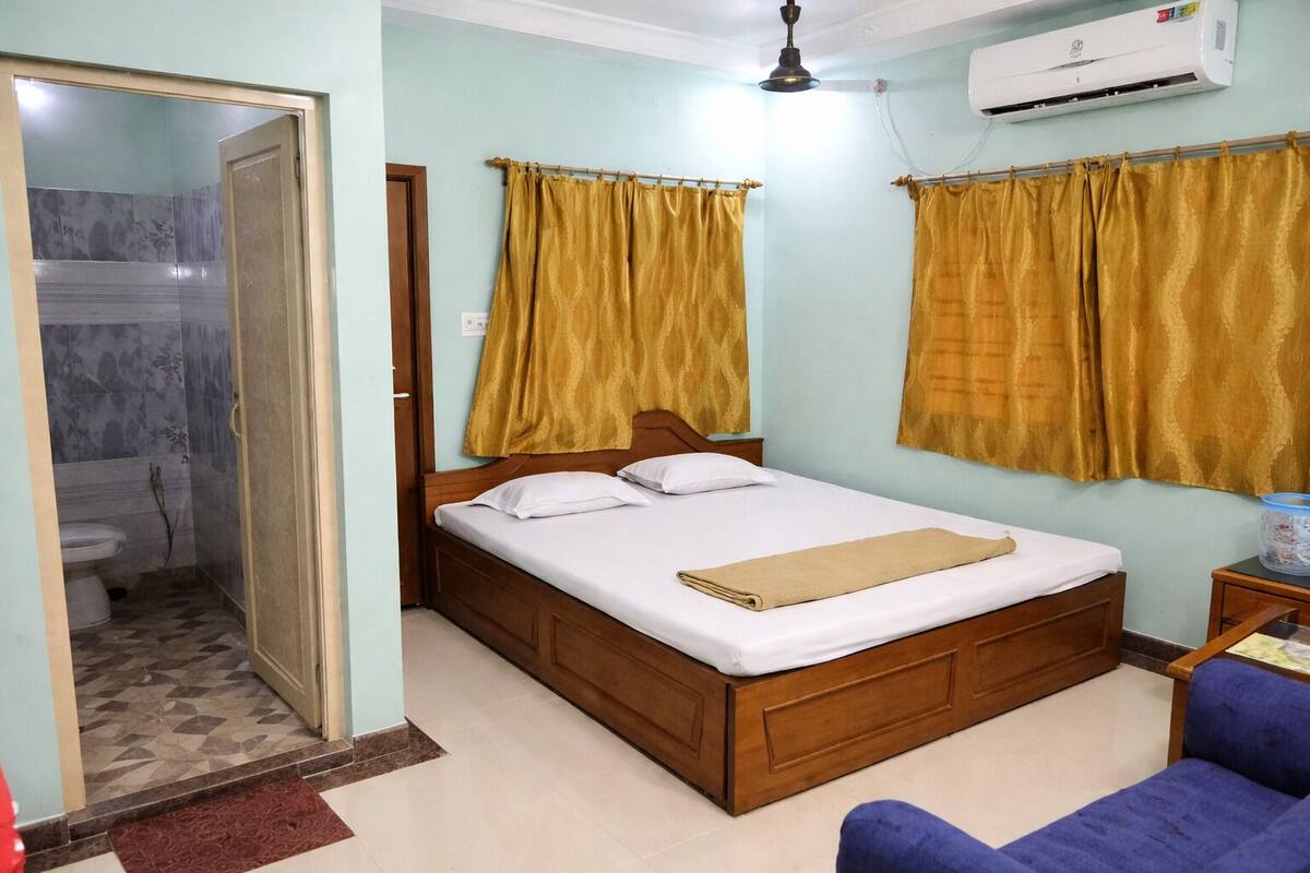 Une chambre avec un lit en bois, une chaise bleue, un climatiseur et une salle de bain équipée de toilettes et d’une douche.
