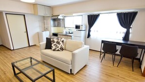 Un espacio habitable compacto con sofá, una mesa de centro de vidrio, una mesa de comedor con sillas, una kitchenette y un ventanal grande con cortinas.