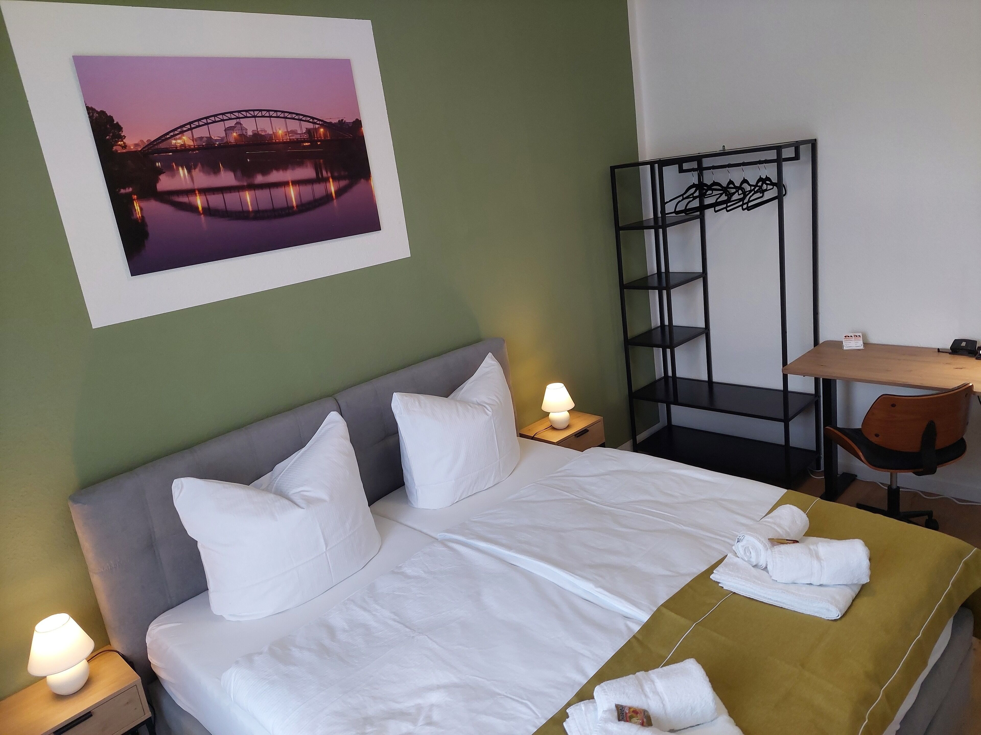 Una cama bien hecha con sábanas blancas, una lámpara de noche y un cuadro con un puente al anochecer.