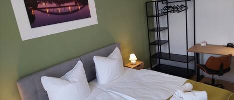 Una cama bien hecha con sábanas blancas, una lámpara de noche y un cuadro con un puente al anochecer.