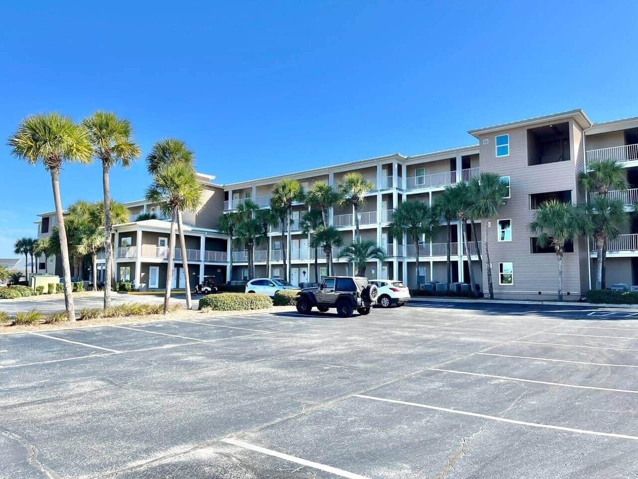 A multi-story hotel with a parking lot, palm trees, and a clear blue sky.