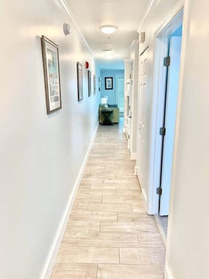 A hallway with framed pictures on the walls, a light-colored wooden floor, and a door on the right.