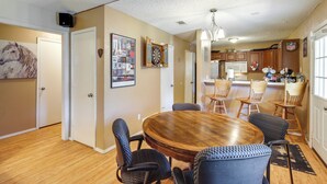 Un coin repas avec une table en bois et six chaises, une cuisine à l’arrière-plan et un tableau encadré au mur.