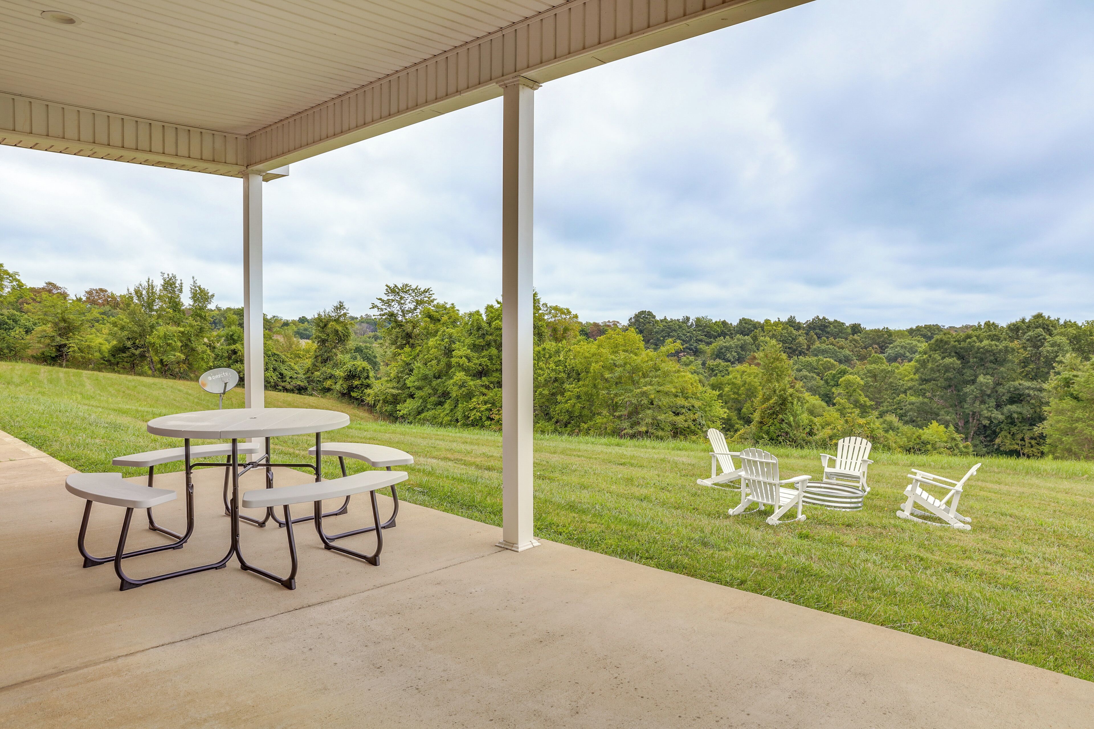 A covered patio with picnic tables and Adirondack chairs, overlooking a grassy area with trees.
