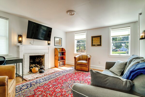 A living room with a fireplace, a television mounted on the wall, a leather armchair, a grey sofa, and a patterned rug. - Charming Victorian Farmhouse on Historic Main St (Trappe)
