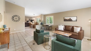 A spacious living area with a brown leather sofa, green armchairs, a wooden coffee table, and a dining area with a chandelier.