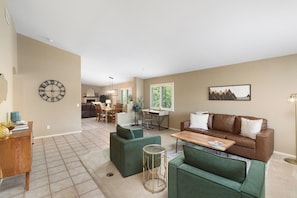A spacious living area with a brown leather sofa, green armchairs, a wooden coffee table, and a dining area with a chandelier.