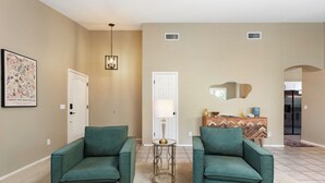 A living room with two green armchairs, a coffee table, a lamp, a white door, and a kitchen area in the background.
