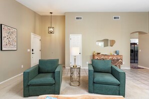 A living room with two green armchairs, a coffee table, a lamp, a white door, and a kitchen area in the background.