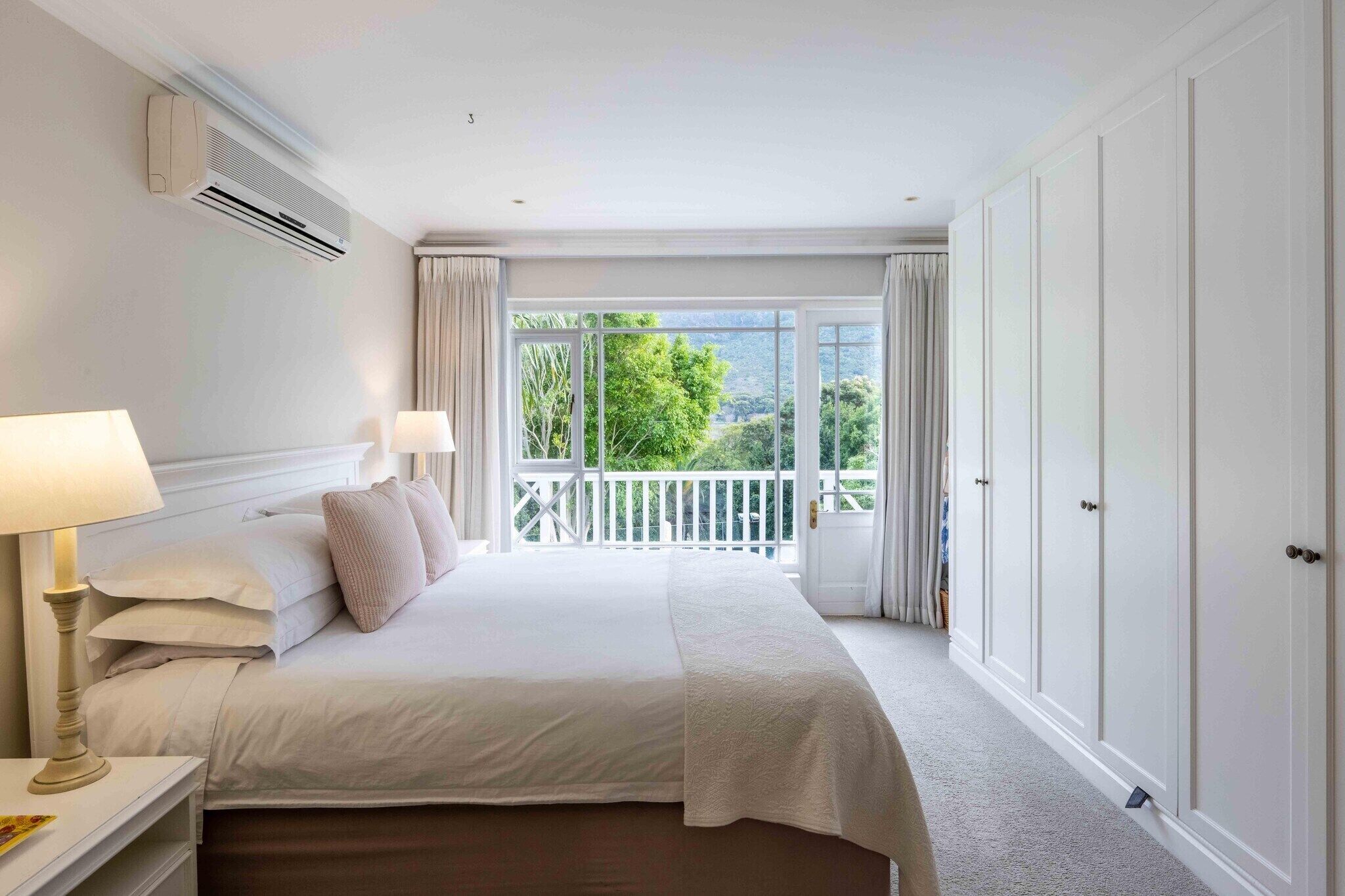 A hotel room with a large bed, two bedside lamps, an air conditioning unit, a balcony with curtains, and a view of greenery.
