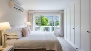 A hotel room with a large bed, two bedside lamps, an air conditioning unit, a balcony with curtains, and a view of greenery.
