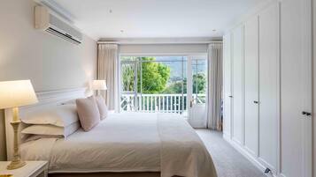 A hotel room with a large bed, two bedside lamps, an air conditioning unit, a balcony with curtains, and a view of greenery.