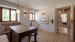 Une pièce spacieuse avec une table à manger en bois, deux chaises et un banc. Il y a deux fenêtres avec des rideaux, un luminaire fixé au mur et un tableau encadré. La pièce est dotée d’un plancher en bois et d’une porte qui donne sur une autre pièce.