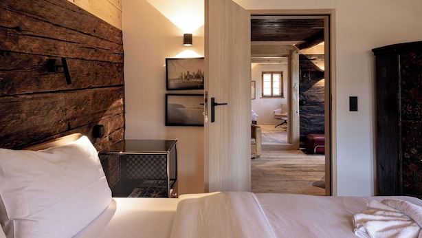 Une chambre avec un plafond en bois, un lit recouvert de draps blancs, un radiateur fixé au mur et une porte donnant sur une autre pièce.