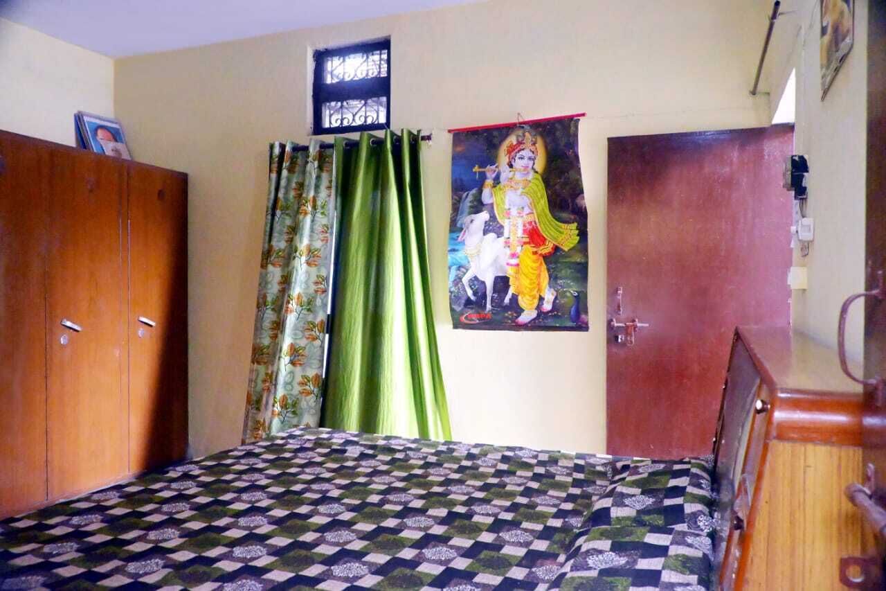 A bedroom with a checkered bedspread, a wooden wardrobe, green curtains, and a religious poster on the wall.