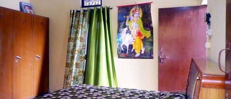 A bedroom with a checkered bedspread, a wooden wardrobe, green curtains, and a religious poster on the wall.