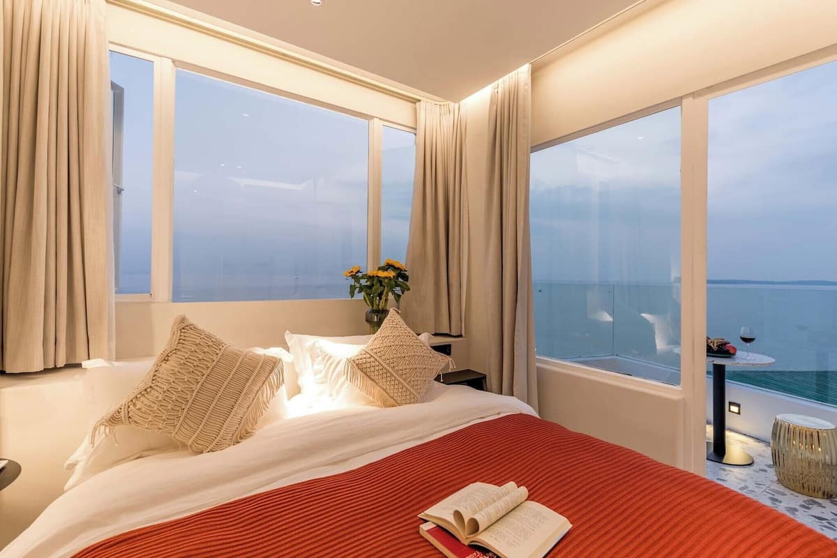 Une chambre d’hôtel avec un grand lit, deux fenêtres avec des rideaux, une petite table avec une boisson, et une vue sur la mer.