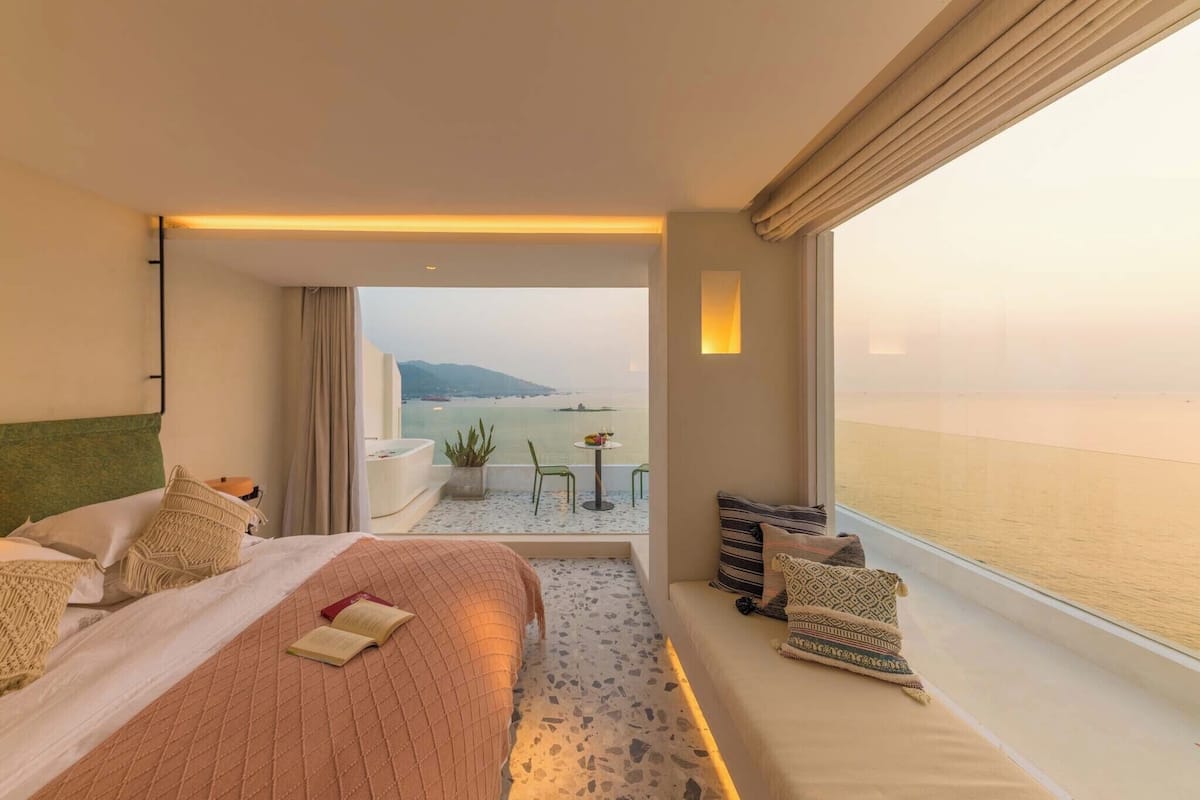 Une chambre d’hôtel avec un lit, une petite table avec une plante, un canapé avec des coussins et une vue sur la mer.