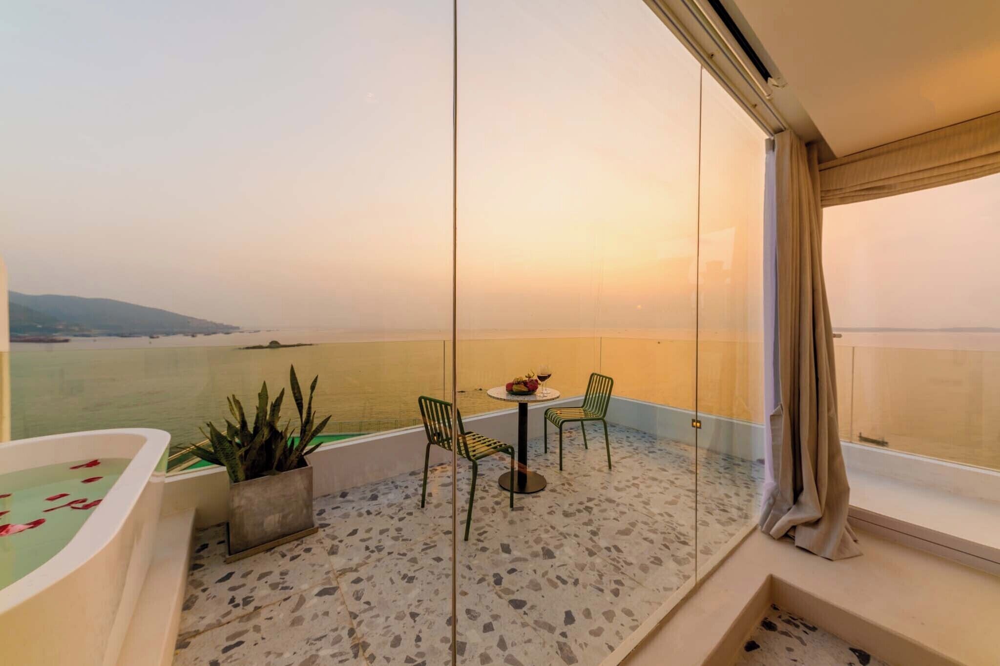 Un balcón con suelo de terrazo, una pequeña mesa con sillas, una planta en maceta y vistas al mar al atardecer.
