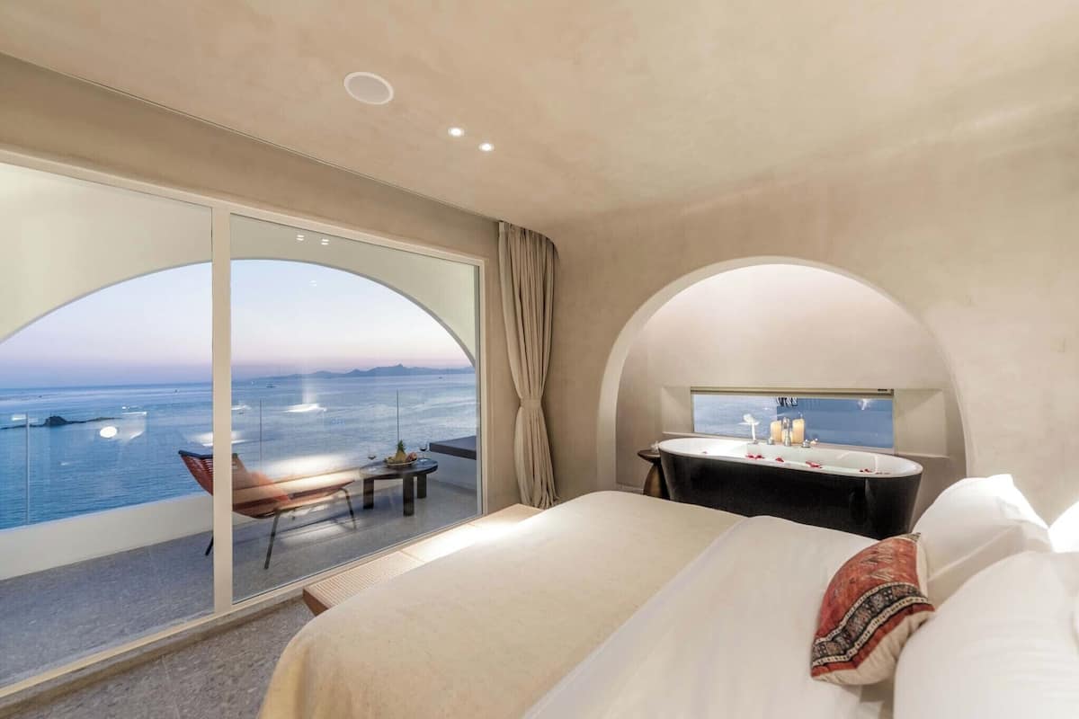 Une chambre d’hôtel moderne dotée d’un grand lit, d’une salle de bain avec baignoire et d’un balcon offrant une vue sur la mer.