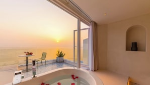 Une pièce avec une grande baignoire remplie d’eau et de pétales de rose, une table avec une bouteille et des verres, une chaise et une plante en pot.